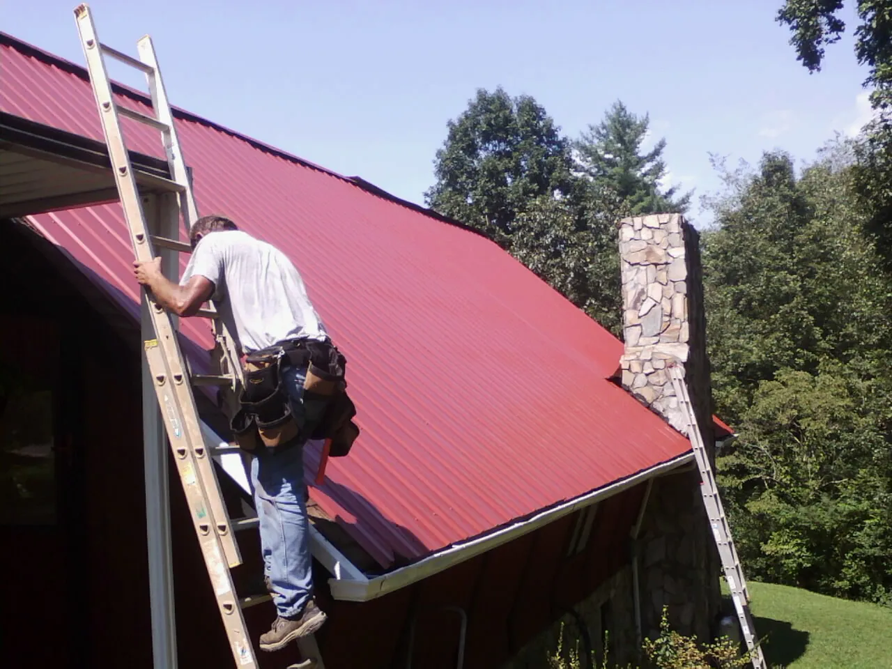 Roofer on a ladder beside a red metal roof and stone chimney — Soffit & Fascia Repair by Roofing Brothers in Franklin Square
